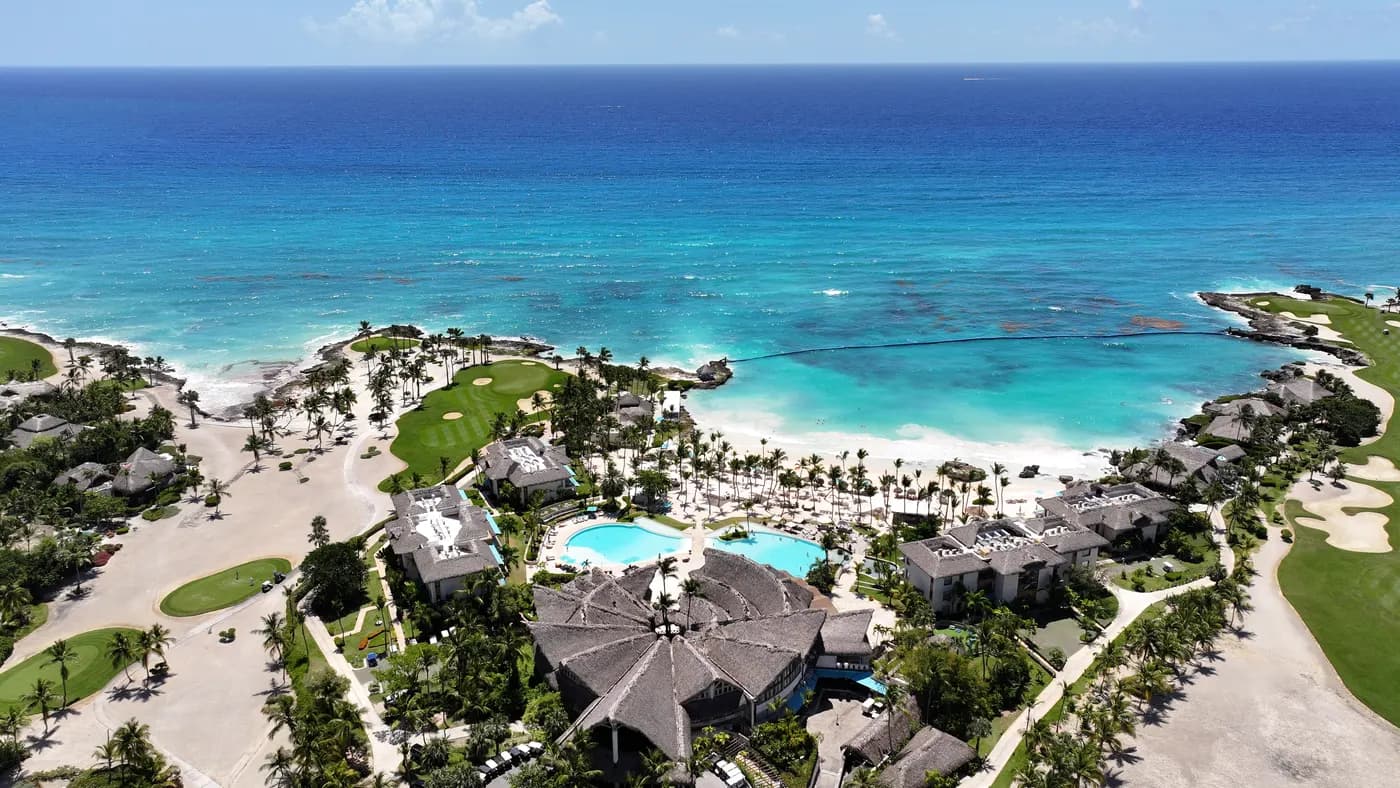 Real aerial photograph of the Caletón cove beach club beside the Punta Espada course, reef water beyond.